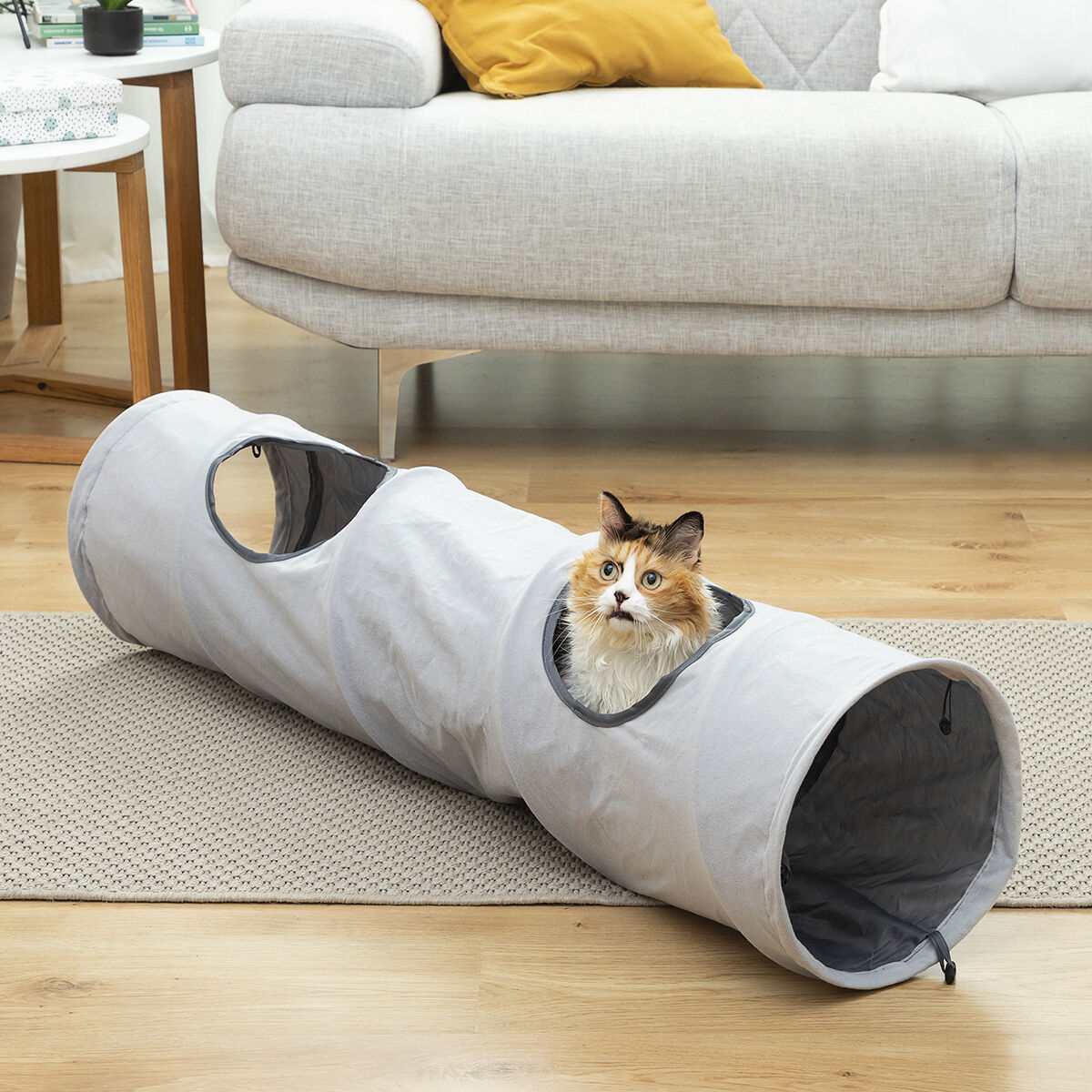 Un gato carey asoma por un túnel gris para gatos sobre una alfombra clara.