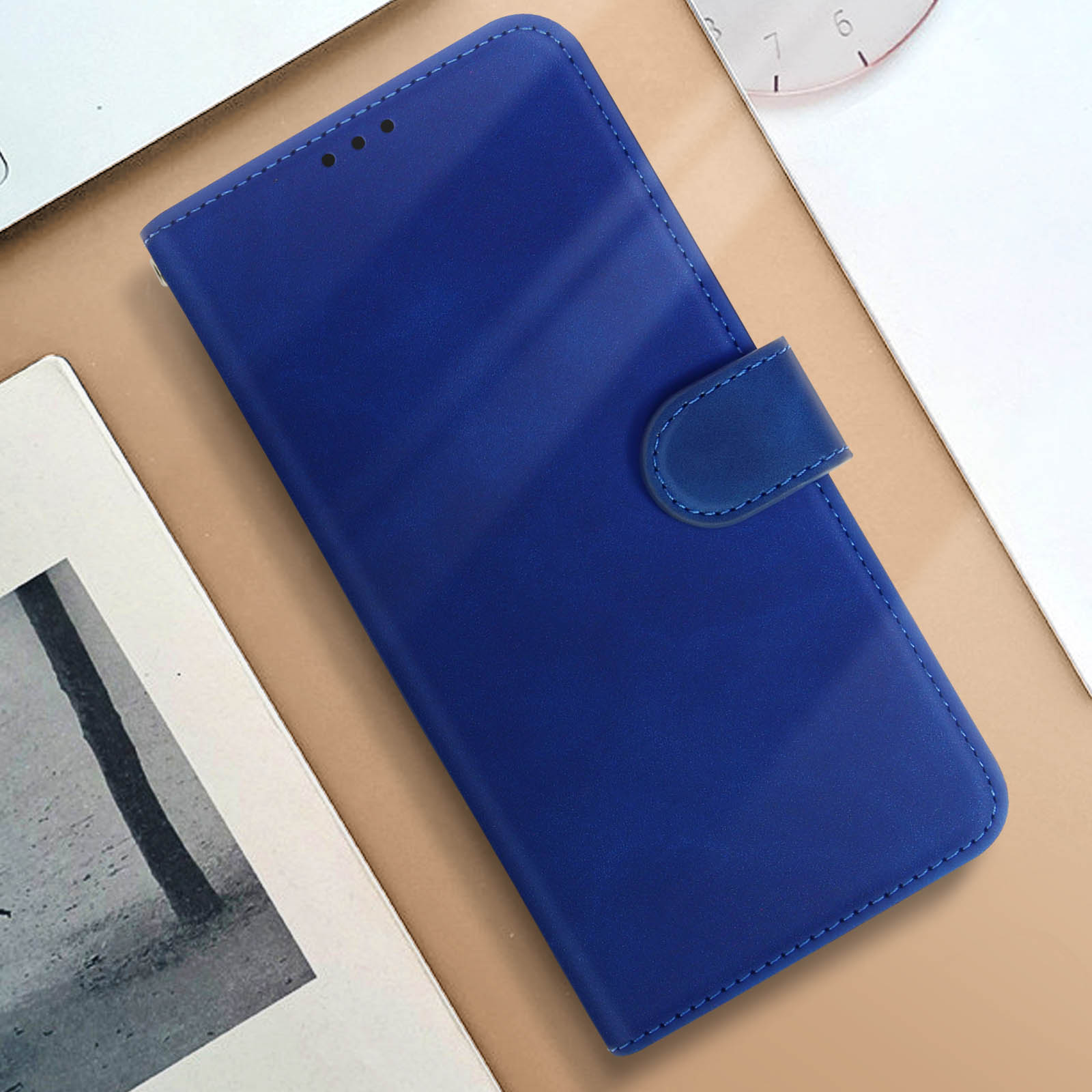 Una custodia per telefono blu, chiusa, con chiusura magnetica, su una superficie beige accanto a un taccuino.