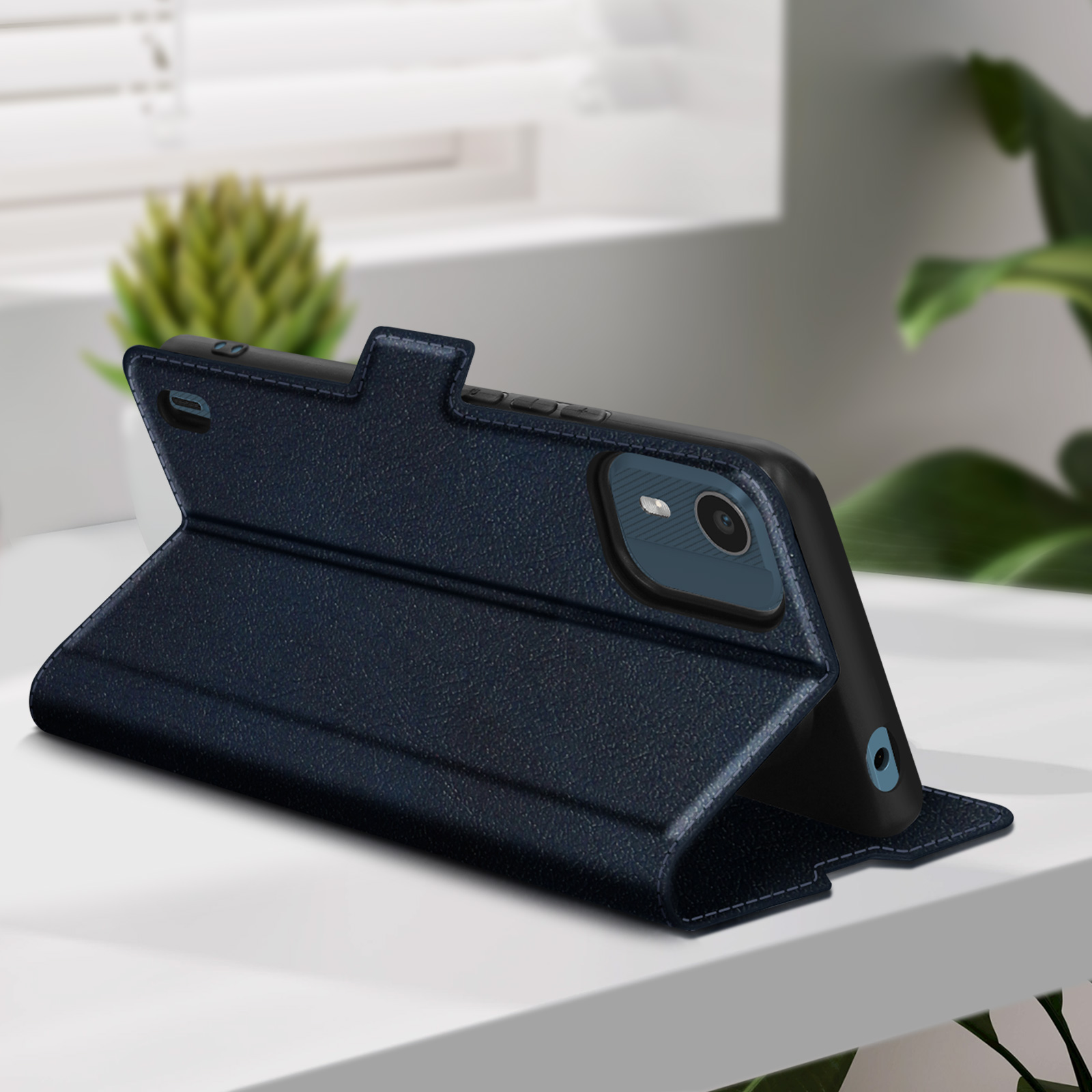 Custodia blu navy con telefono. Custodia aperta, telefono visibile. Tavolo bianco e pianta verde sullo sfondo.