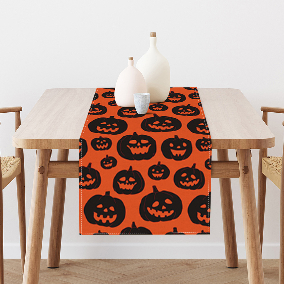 Halloween-Tischläufer mit Kürbisgesichtern auf orangefarbenem Hintergrund, zwei Vasen und einer Tasse.