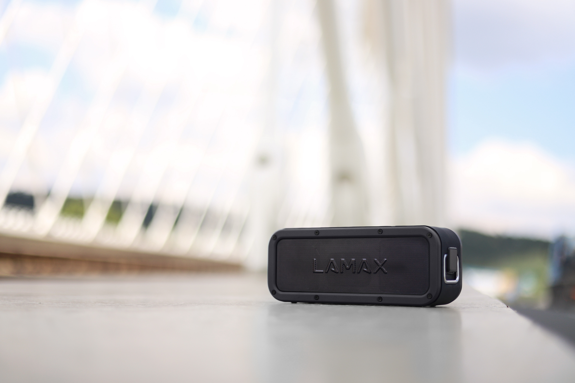 Czarny głośnik LAMAX spoczywający na powierzchni, z rozmytym mostem w tle.