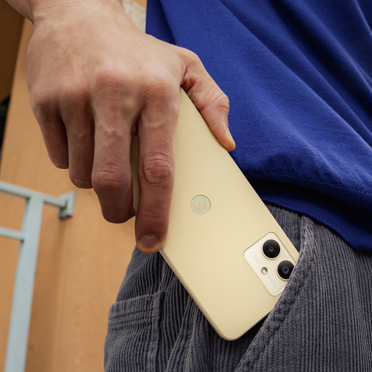 Mano sosteniendo un smartphone beige, parcialmente en un bolsillo. Camisa azul.
