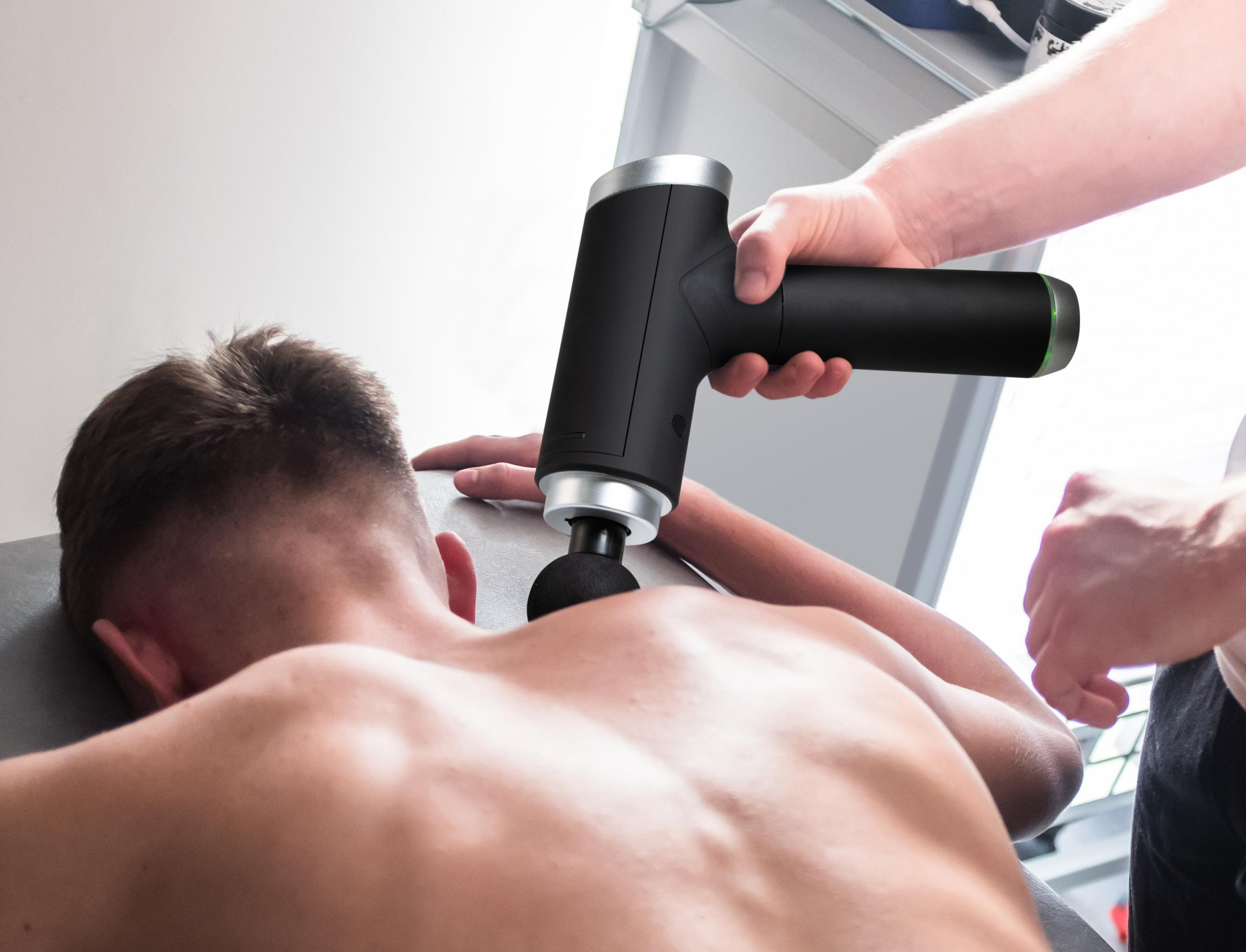 Eine Person benutzt eine schwarze Massagepistole auf dem Rücken einer anderen Person.