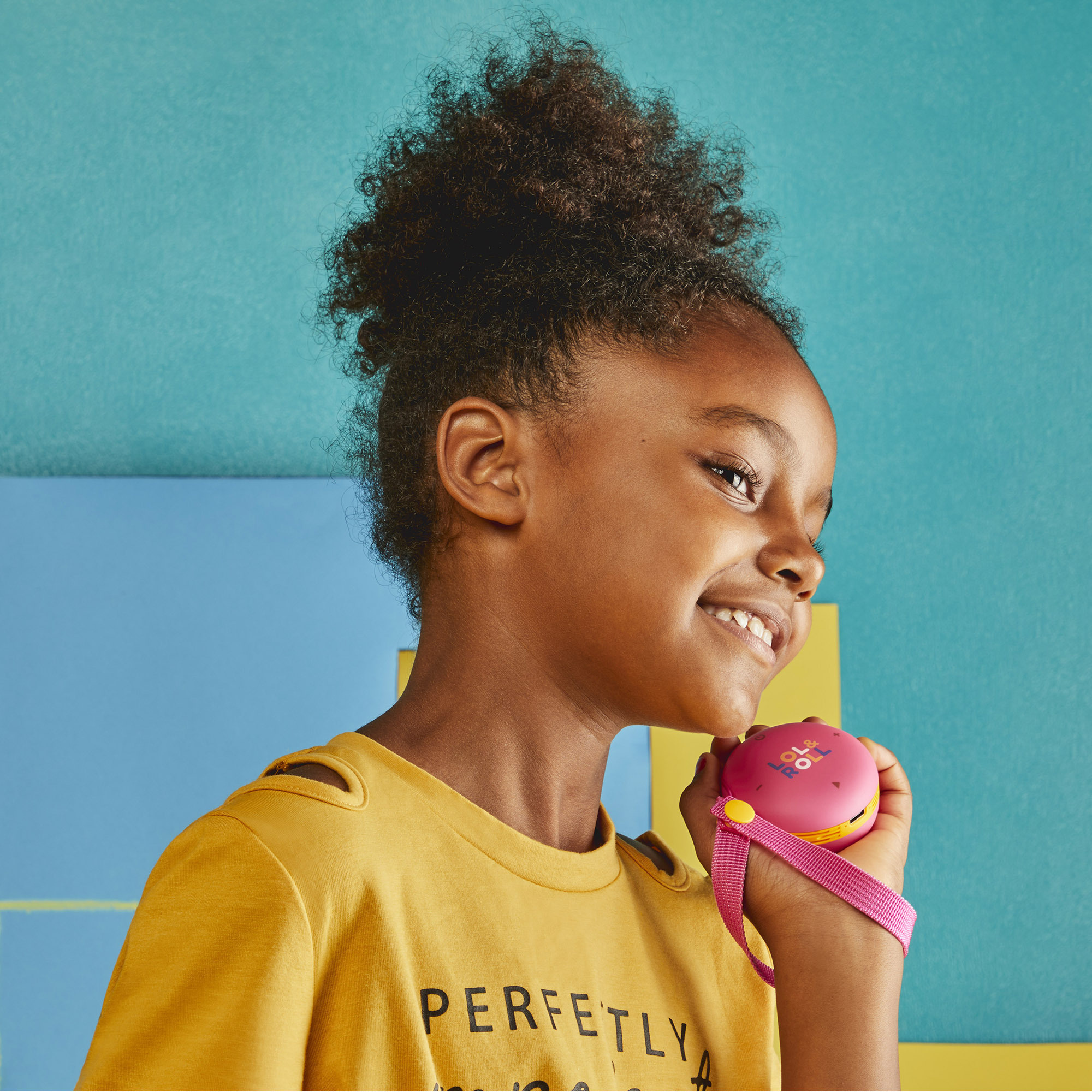 Młoda dziewczyna uśmiecha się, trzymając różowe urządzenie z różowym paskiem na tle niebiesko-żółtym.