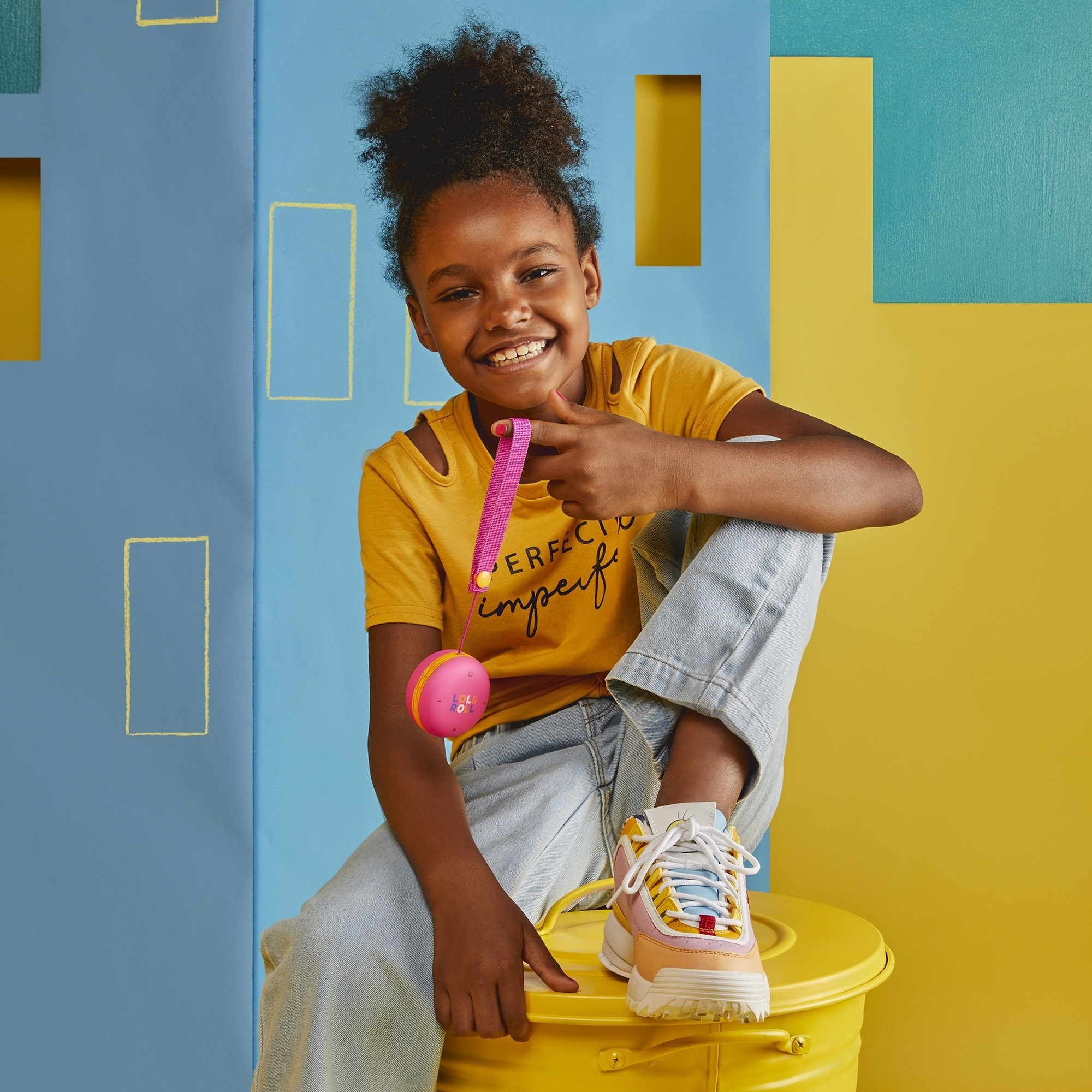 Una niña sonriente sosteniendo un juguete rosa, sentada en un bote de basura amarillo.