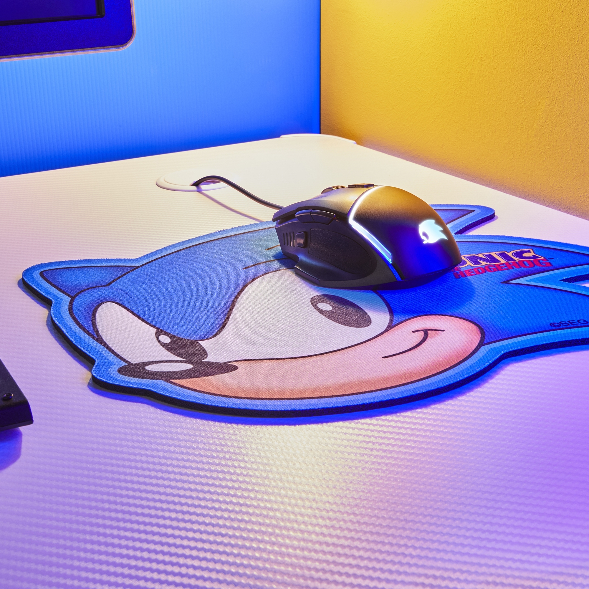 Maus auf Sonic-Mauspad. Blauer Hintergrund, weißer Schreibtisch, gelbe Wand.