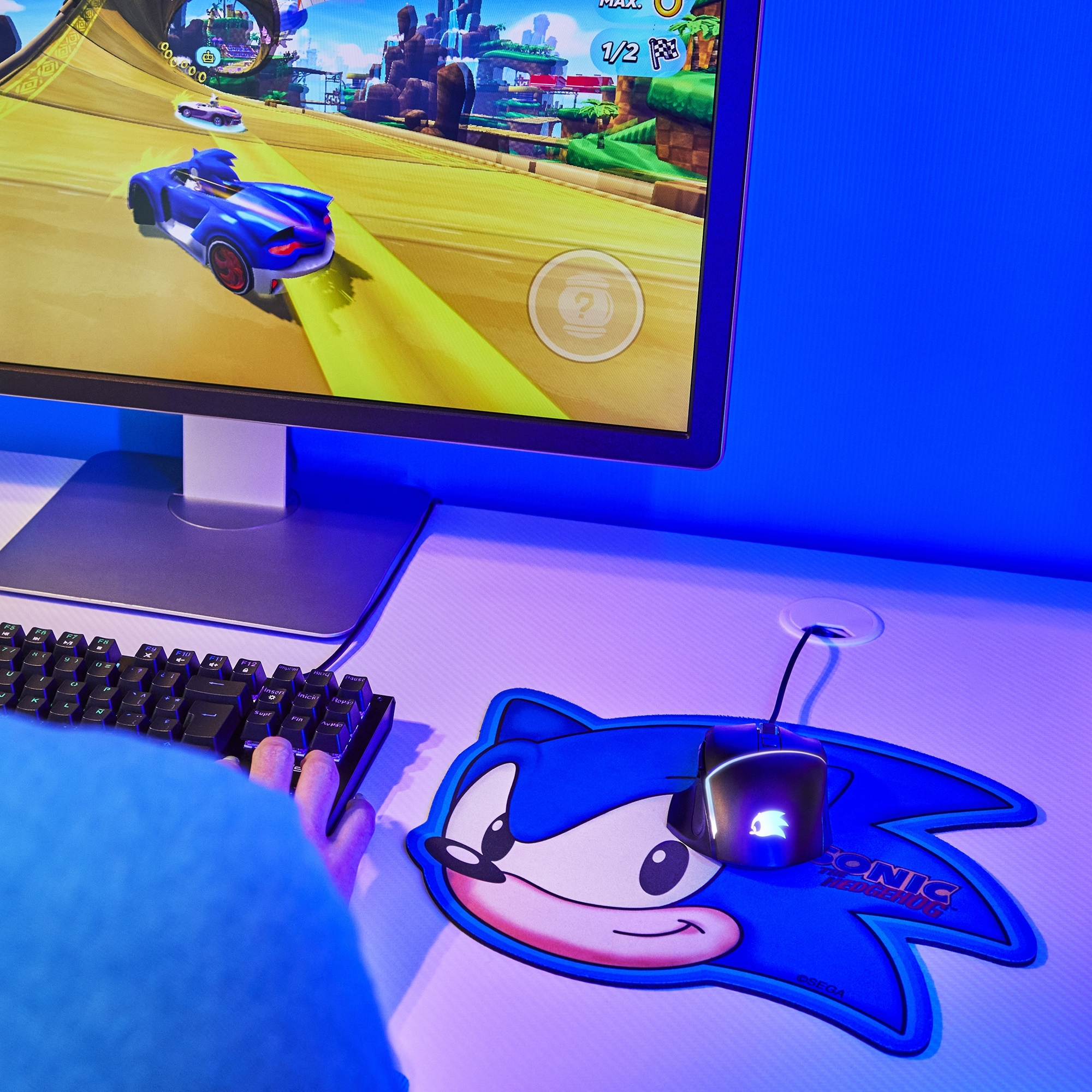 Una configuración de juego con alfombrilla Sonic azul, teclado, ratón y monitor mostrando un juego de carreras.