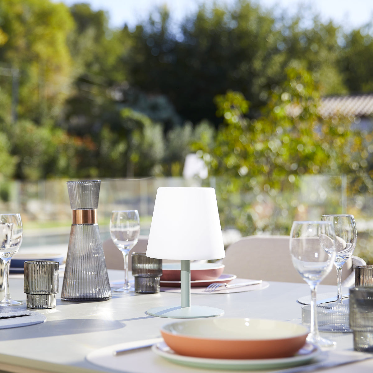 Ein Tisch mit weißer Lampe, Gläsern, Tellern und anderen Gegenständen auf einer Terrasse mit Bäumen im Hintergrund.