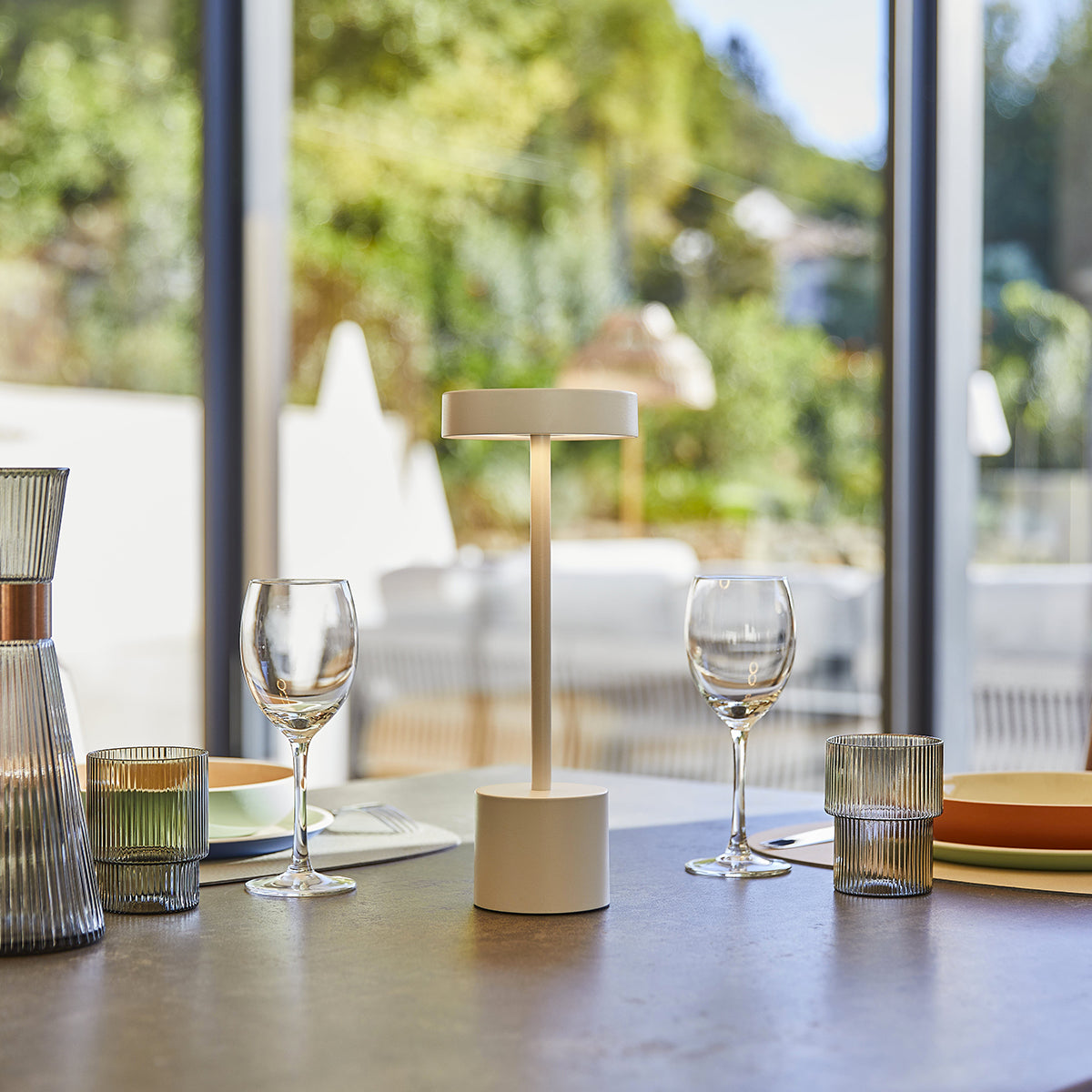 Eine weiße und beige Lampe auf einem Tisch mit Gläsern und einem Fensterblick ins Grüne.