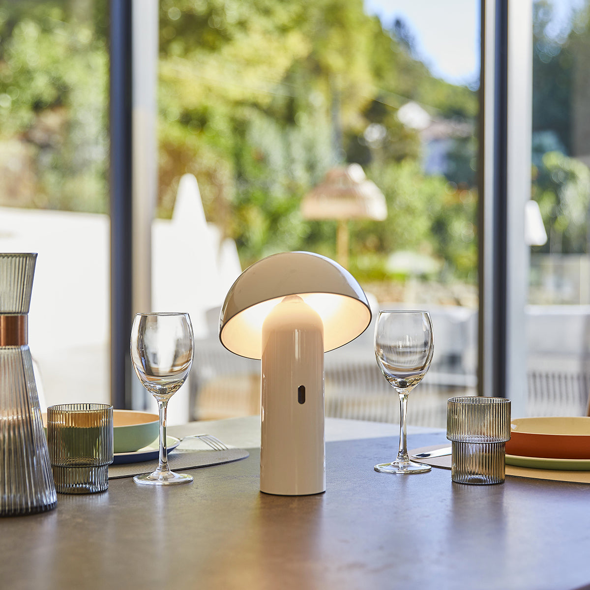Eine weiße Pilzlampe auf einem Tisch mit Gläsern, einer Kerze und Grün im Hintergrund.