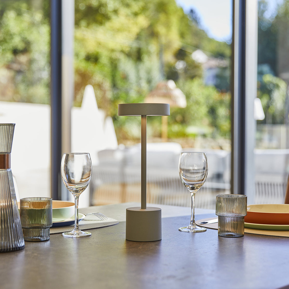 Moderne Lampe auf einem Tisch mit Weingläsern, einem grünen Kerzenhalter und einem Fenster.
