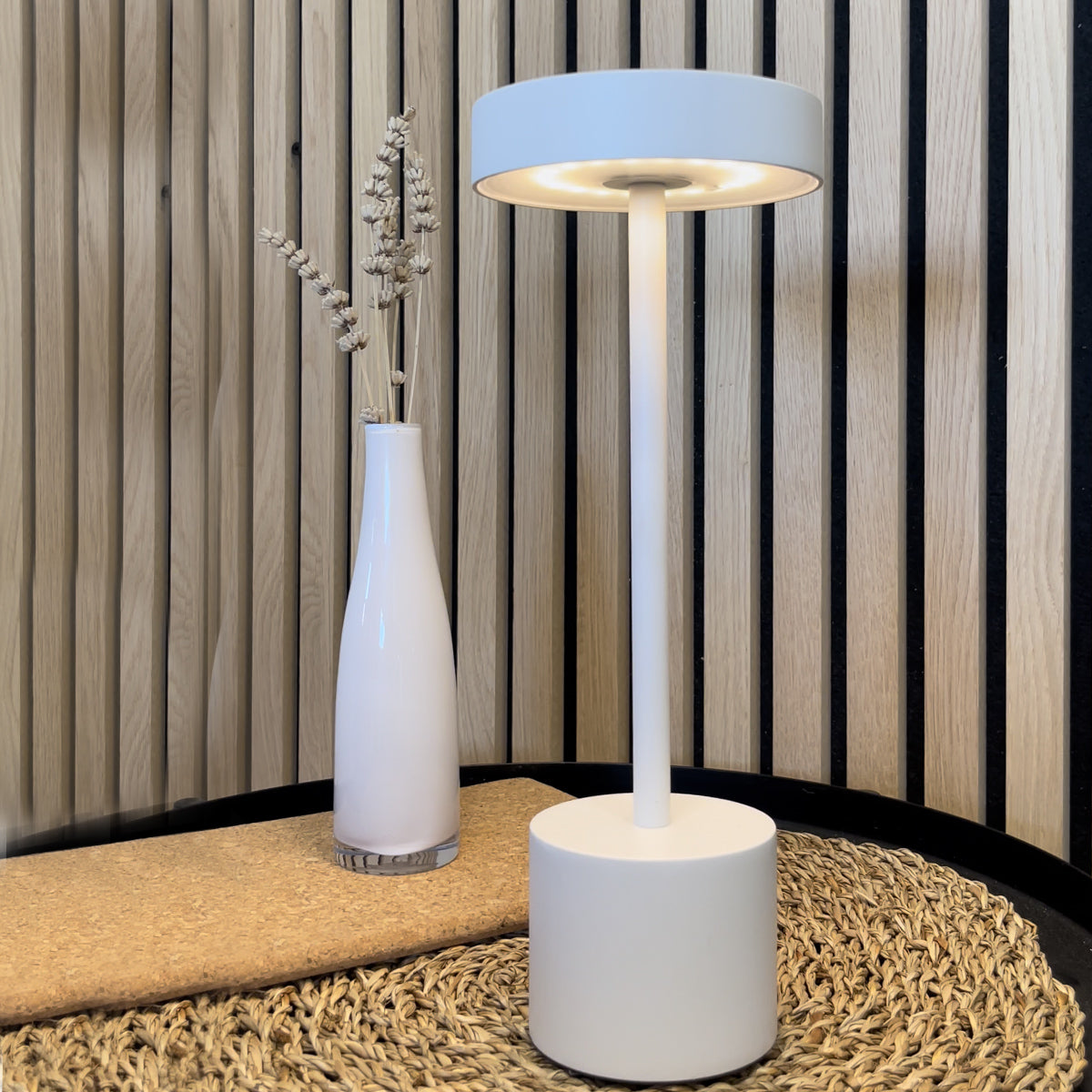 Eine weiße Tischlampe mit zylindrischem Sockel, neben einer Vase mit Trockenblumen.