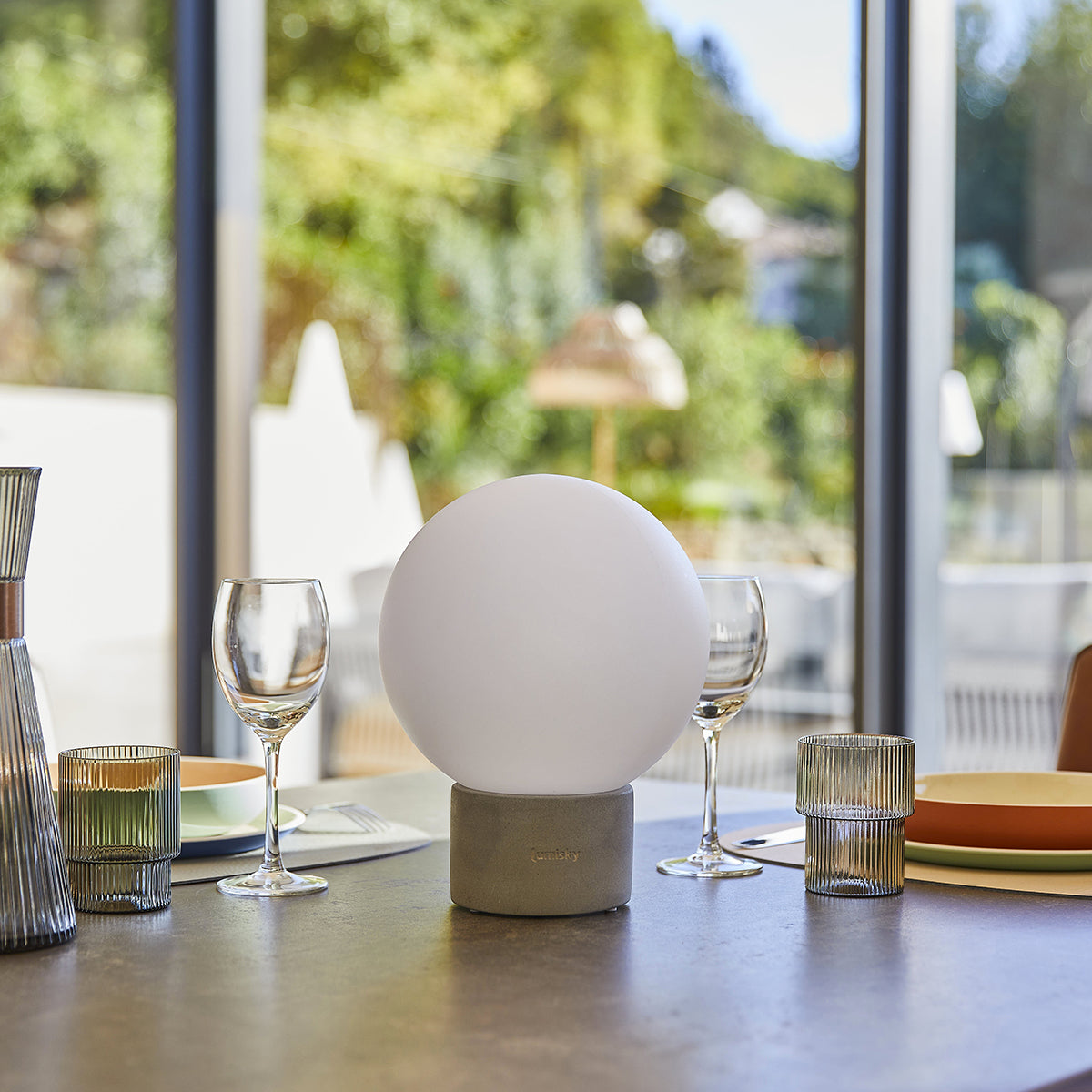 Eine weiße kugelförmige Lampe auf grauem Sockel, Weingläser und Dekogläser. Fenster und Garten sind sichtbar.
