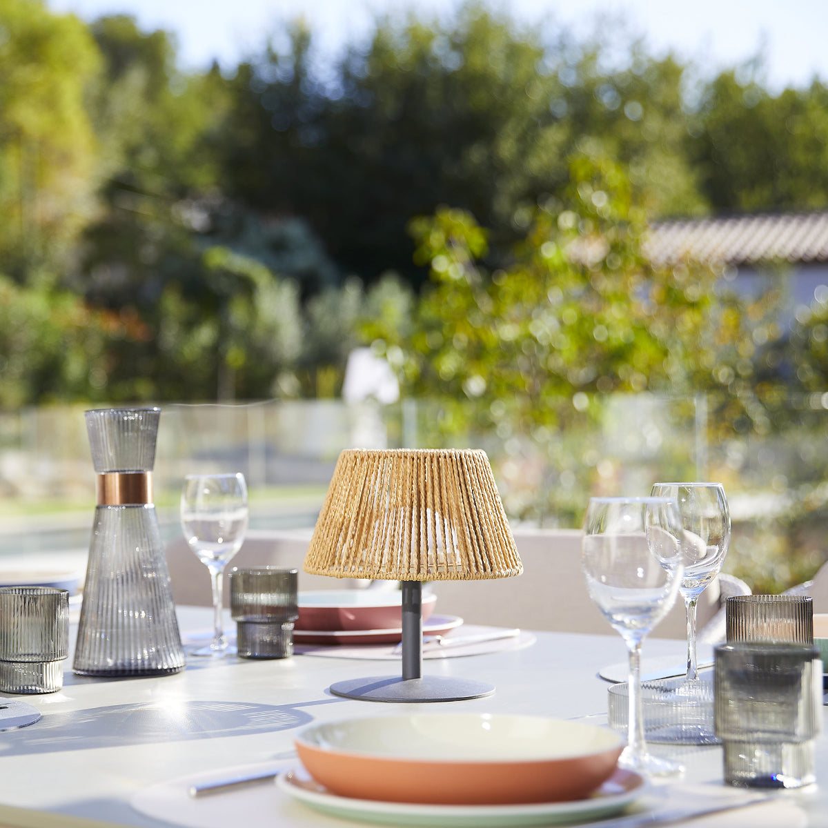 Tisch mit Lampe, Gläsern und Geschirr. Im Hintergrund Bäume und Sonnenlicht.
