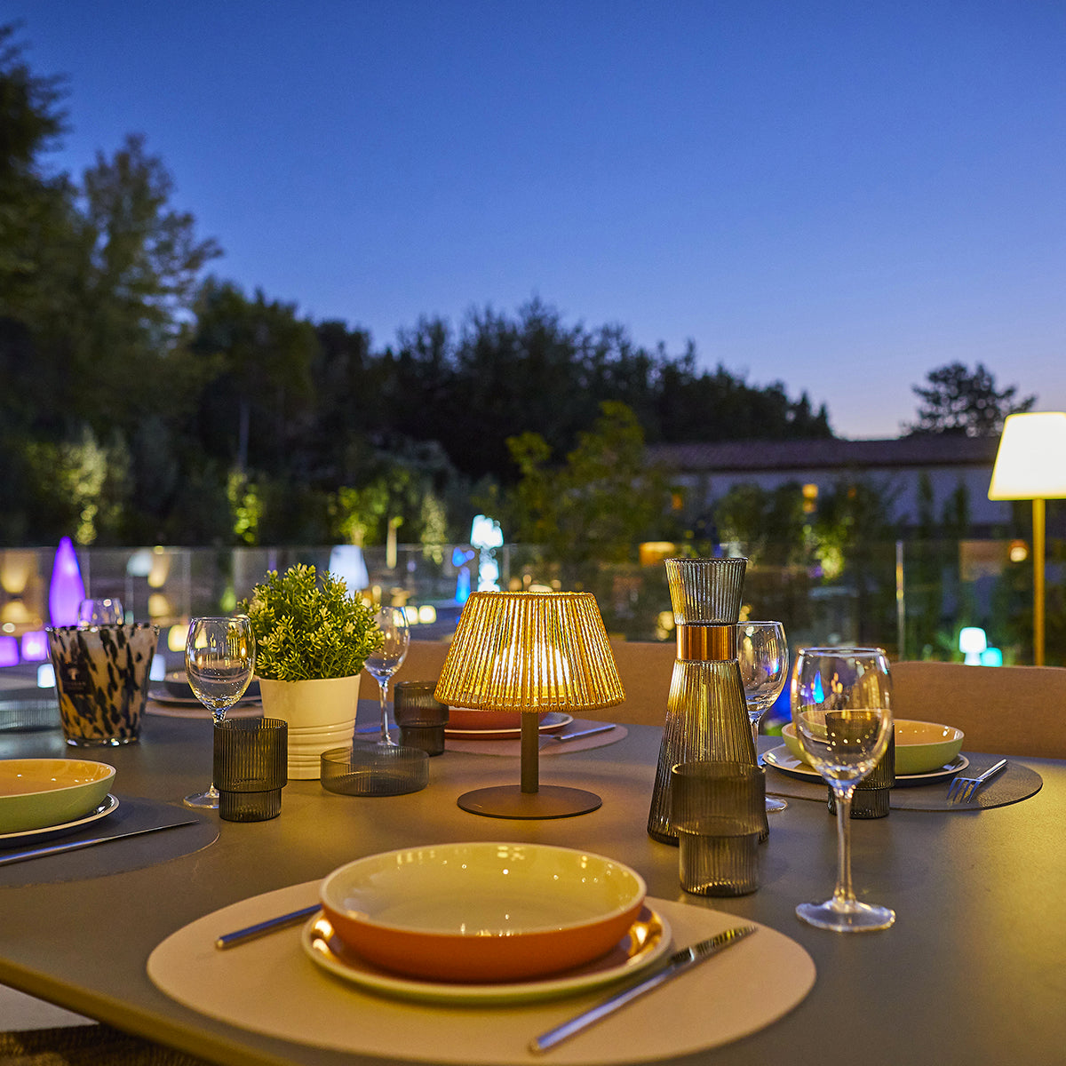 Outdoor-Esstisch für eine Nacht; beleuchtete Tischlampe, Gläser, Teller und Bäume im Hintergrund.