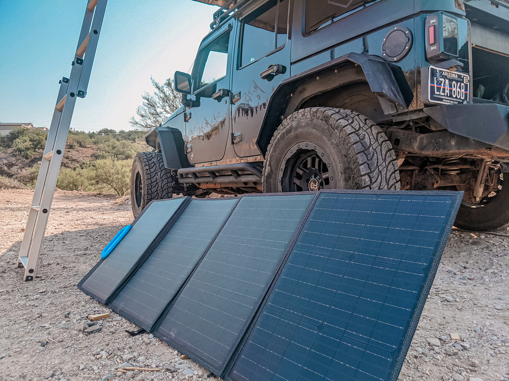 Een zwarte Jeep met zonnepanelen en een ladder op een onverharde weg in de zon.