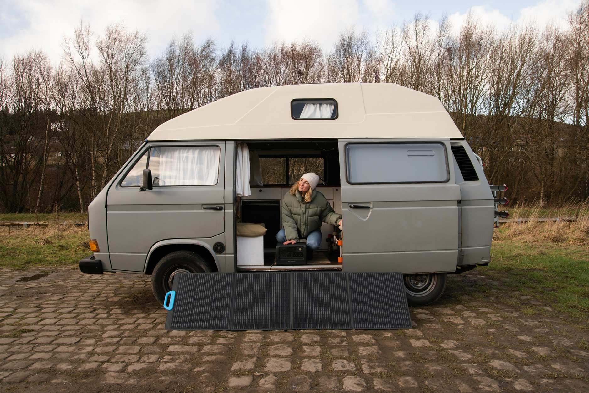 Vrouw in een grijze camper. Deur open, een zonnepaneel uitgevouwen op de grond.