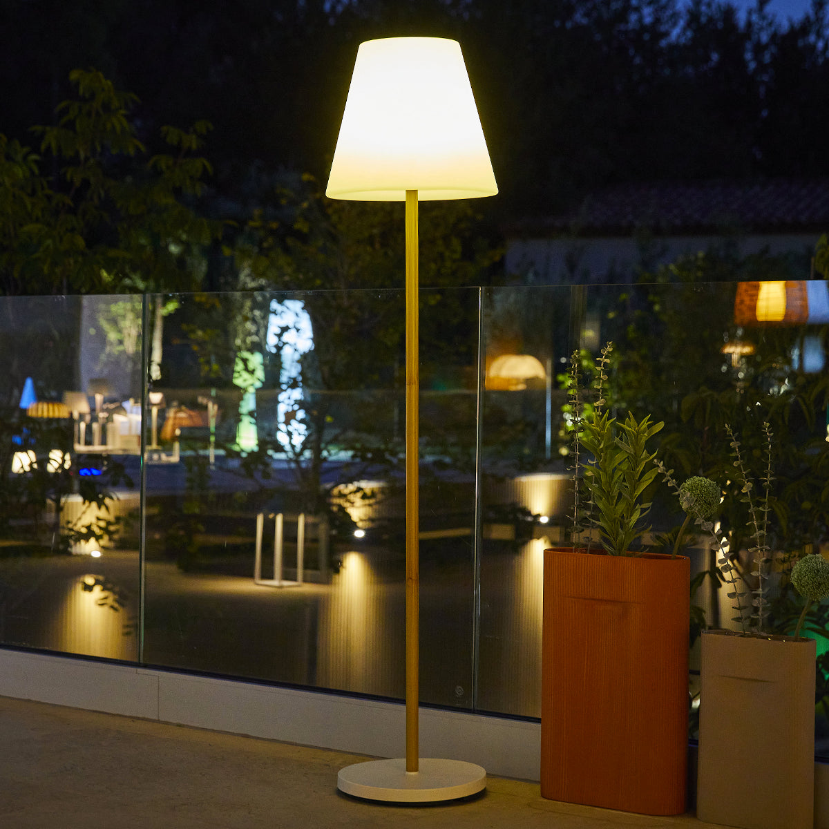 Stehende Stehlampe mit weißem Lampenschirm, Holzstange und weißem Sockel. Dunkler Hintergrund.