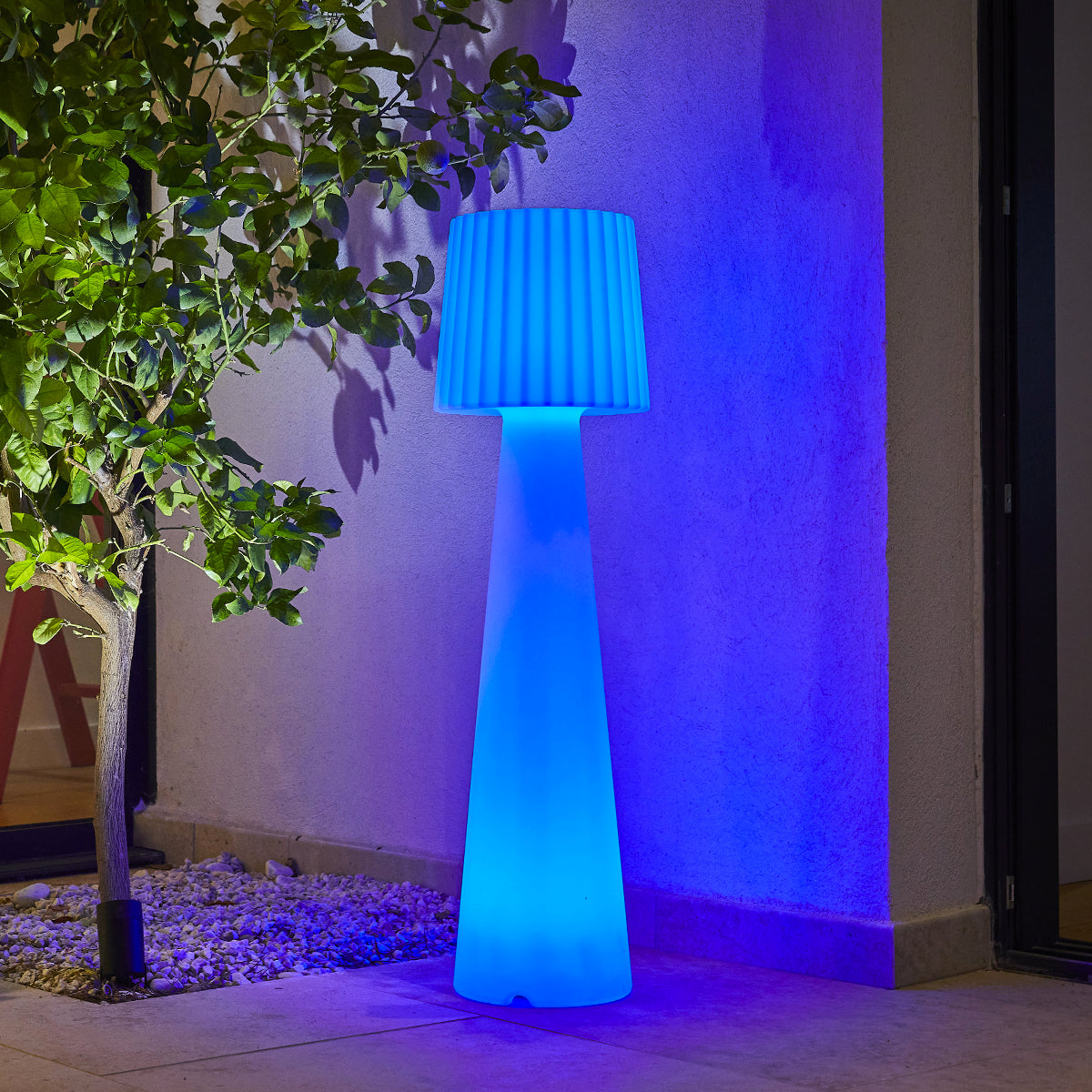 Lampada blu da esterno con base alta e paralume, vicino a un albero e un muro.