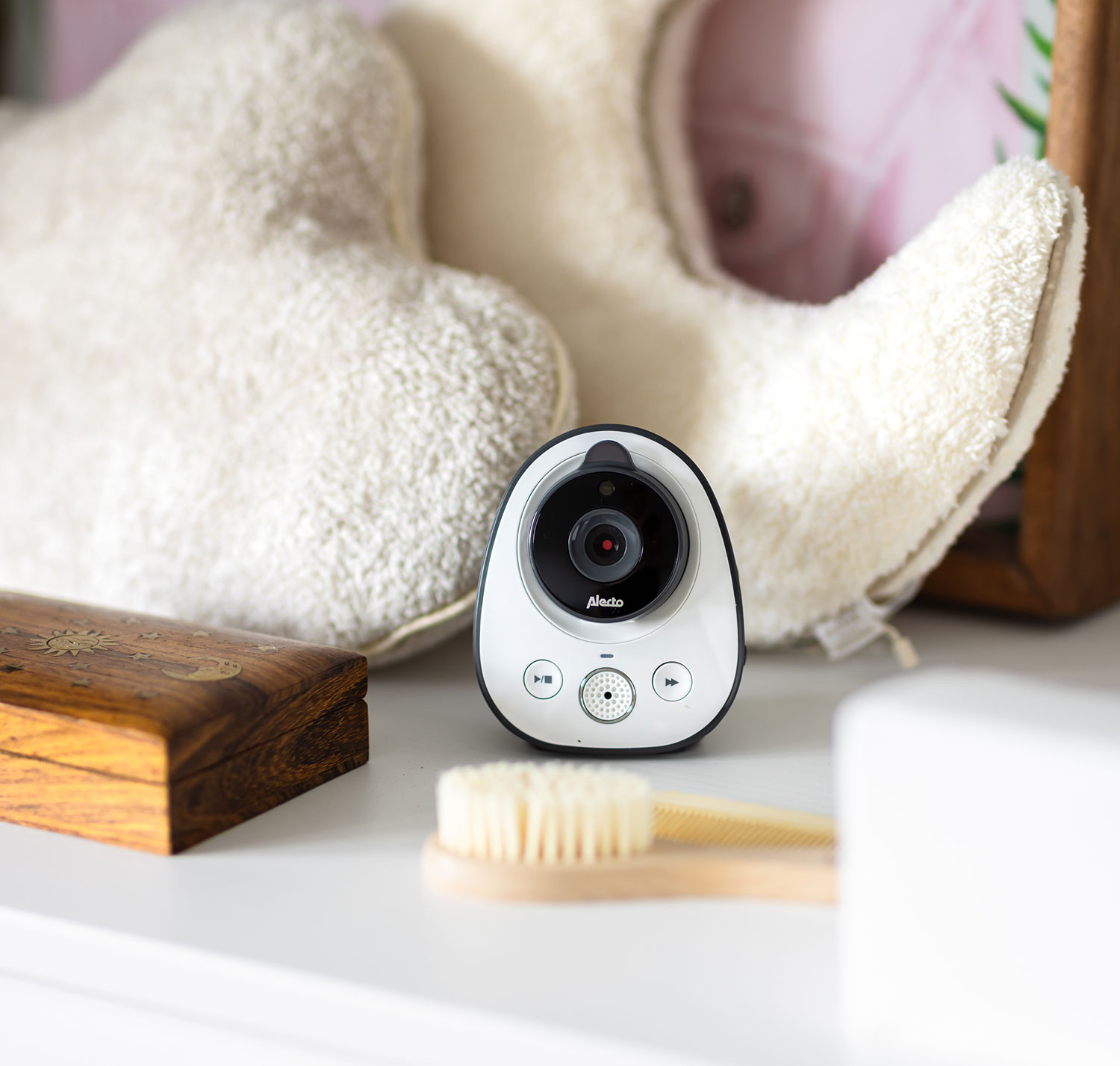 Weißer Babyphone mit Kamera, auf einem Regal platziert. Es befindet sich in der Nähe einer Bürste und eines Plüschtiers.