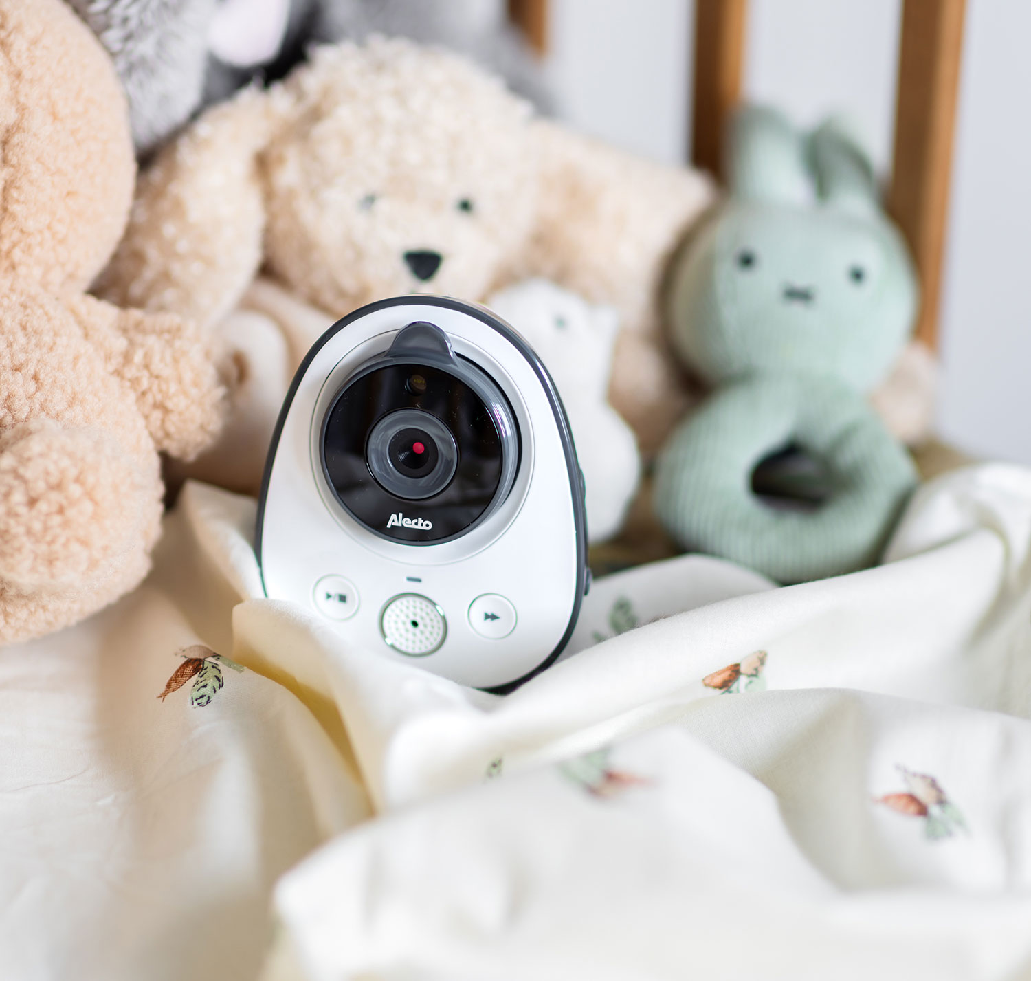 Babyphone auf einem Bett mit Spielzeug. Die Kamera hat ein Objektiv und einen Einschaltknopf.