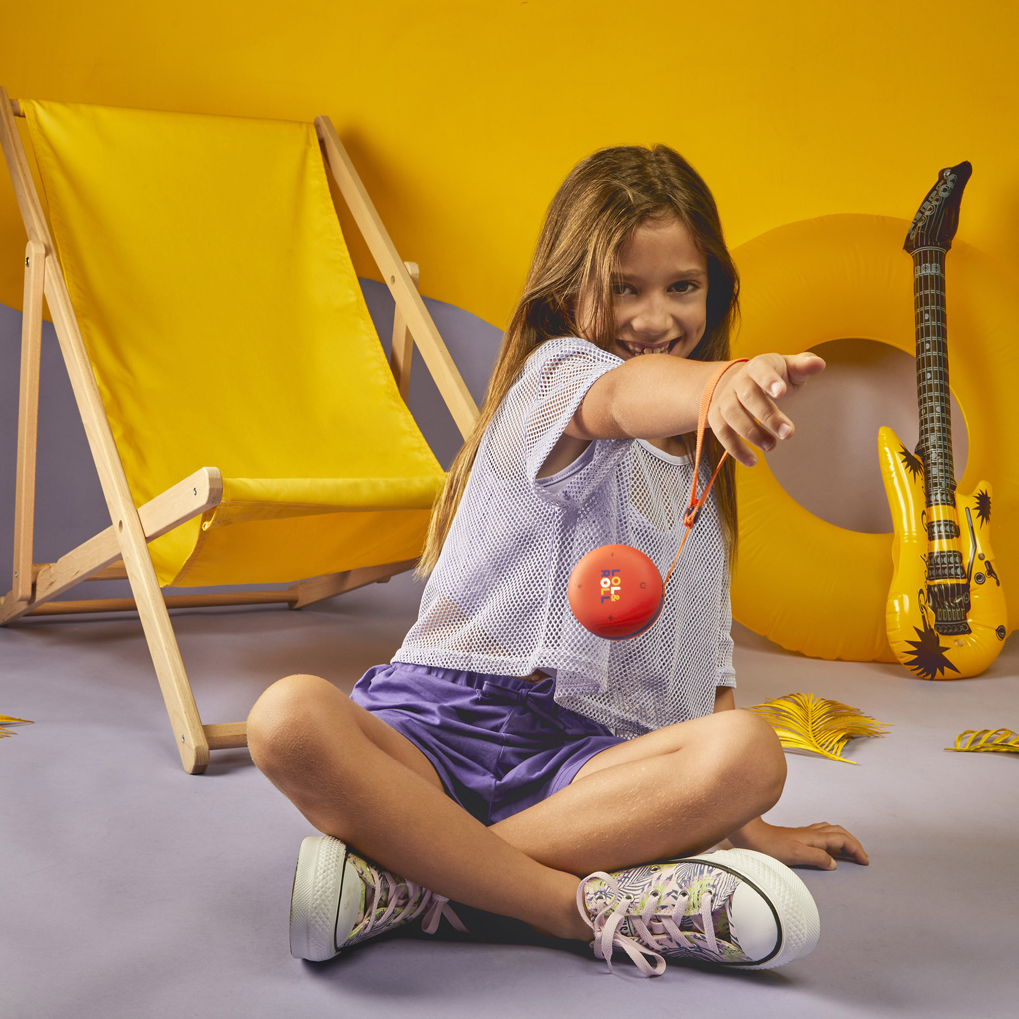 Dziewczyna uśmiecha się, trzymając czerwoną zabawkę, siedząc przed żółtym tłem z akcesoriami plażowymi.