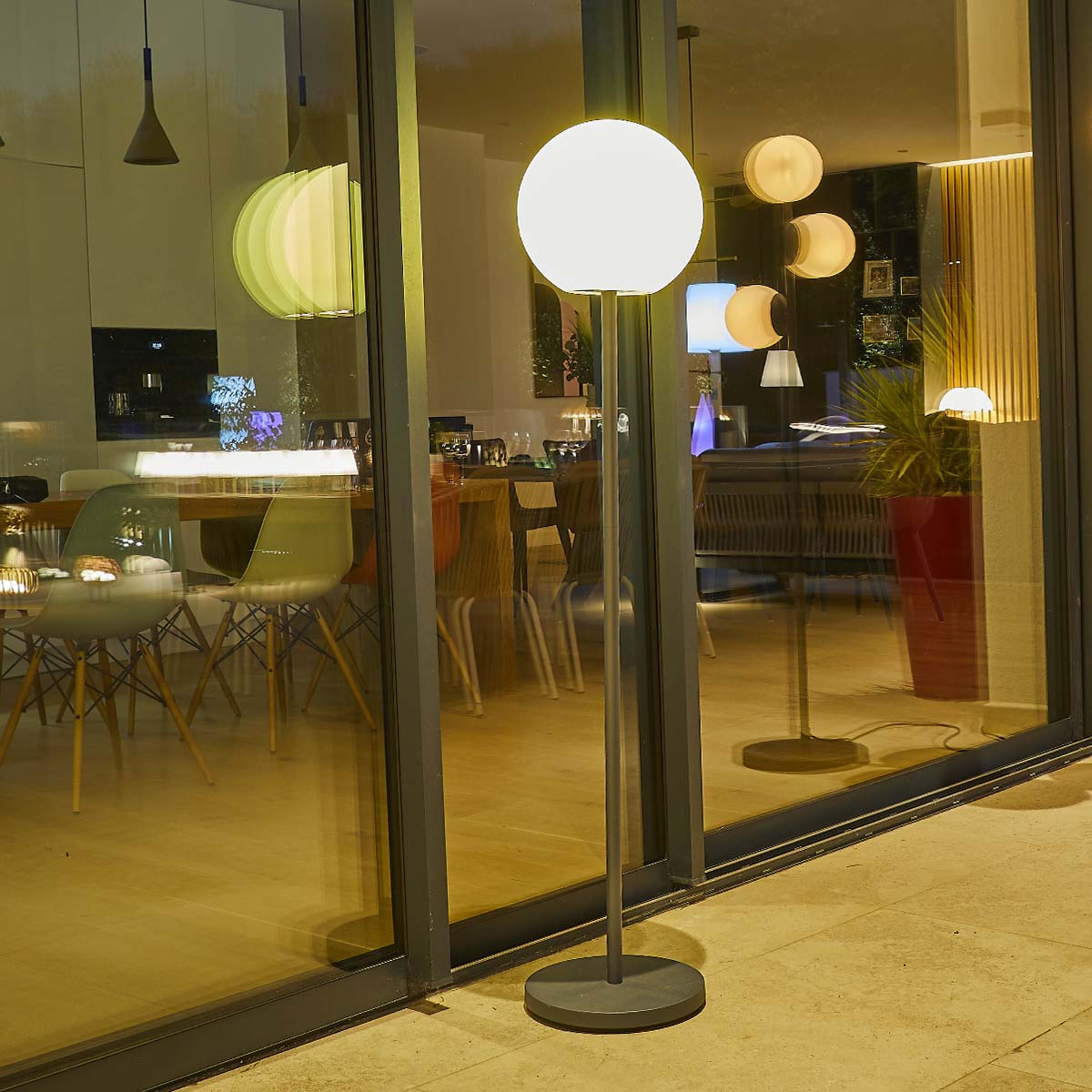 Esterno di casa moderna con una grande lampada da terra che illumina una zona pranzo attraverso una parete di vetro.