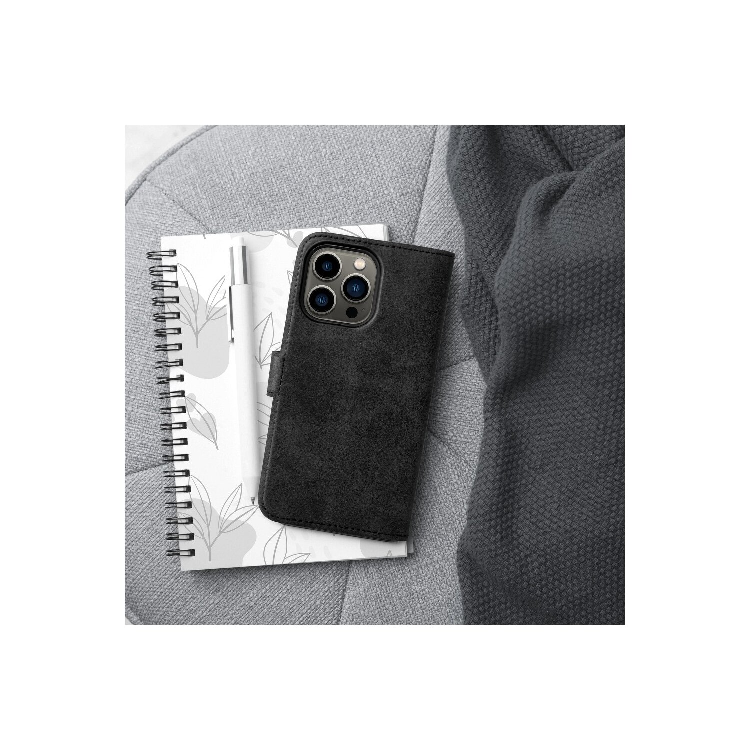 Eine schwarze Leder-Handyhülle mit einem Notizbuch und einem Stift, die auf einer grauen gepolsterten Oberfläche liegen.