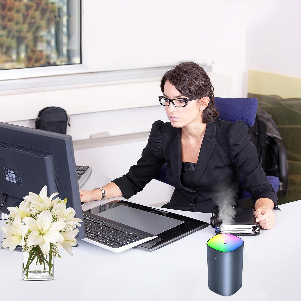 Eine Frau im schwarzen Anzug arbeitet an einem Schreibtisch mit Computer, Blumen und Luftbefeuchter.