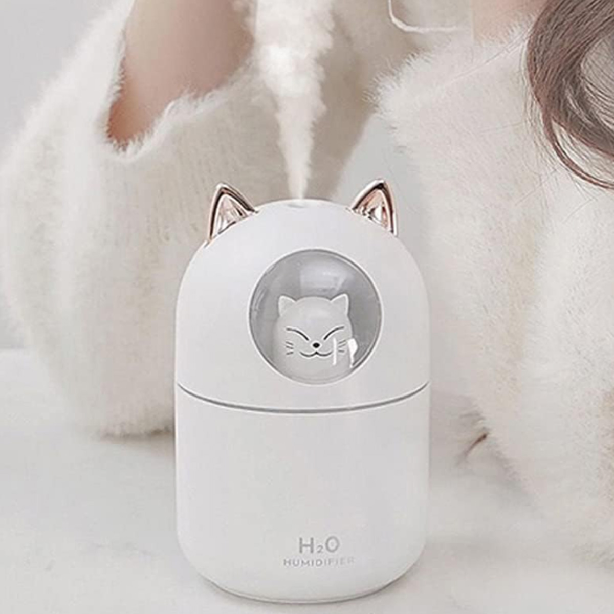 Ein weißer, katzenförmiger Luftbefeuchter mit Ohren, einer Katzen-Gesichtsöffnung und dampfartigem Nebel, drinnen, auf hellem Hintergrund.