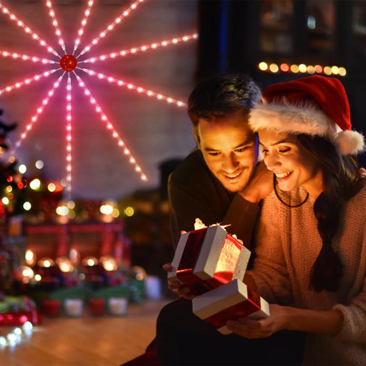 Ein Paar öffnet Geschenke neben einem Weihnachtsbaum mit roter Lichterkette.
