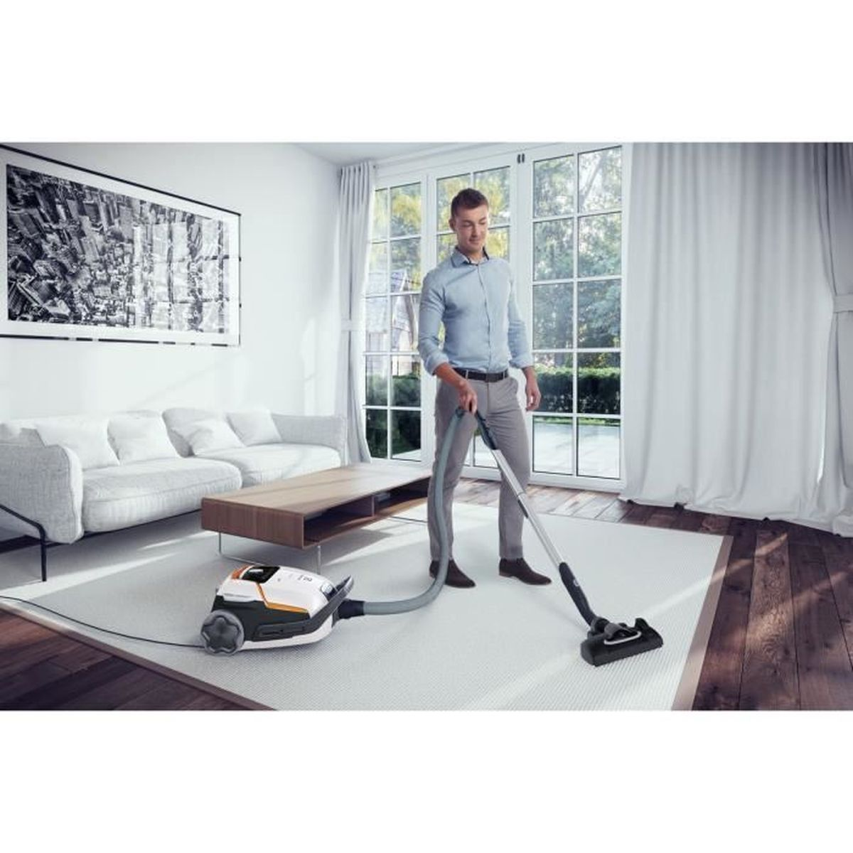 Mężczyzna odkurzający biały dywan odkurzaczem w salonie z dużymi oknami.