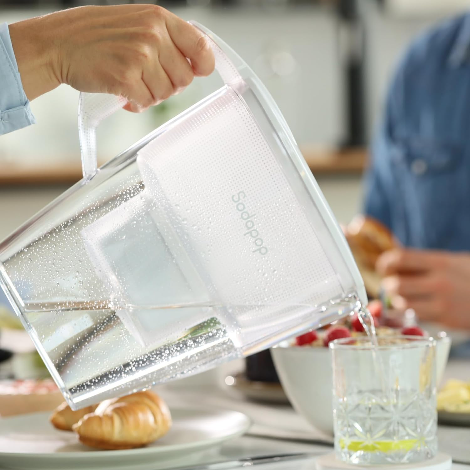 Ein Krug gießt Wasser in ein Glas. Croissant und Essen auf einem Tisch. Eine Person im Hintergrund.
