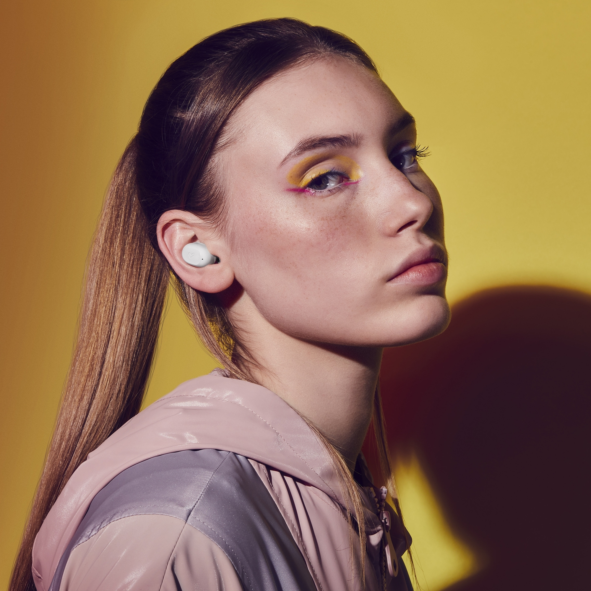 Mujer con cola de caballo usando auriculares, chaqueta rosa y gris, y maquillaje de ojos amarillo vibrante sobre un fondo amarillo.