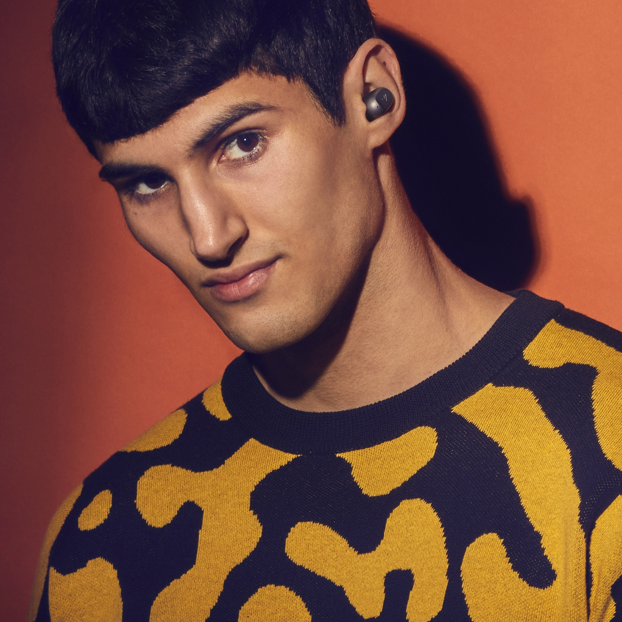 Un hombre con un suéter amarillo estampado sobre un fondo negro. Tiene auriculares en las orejas.