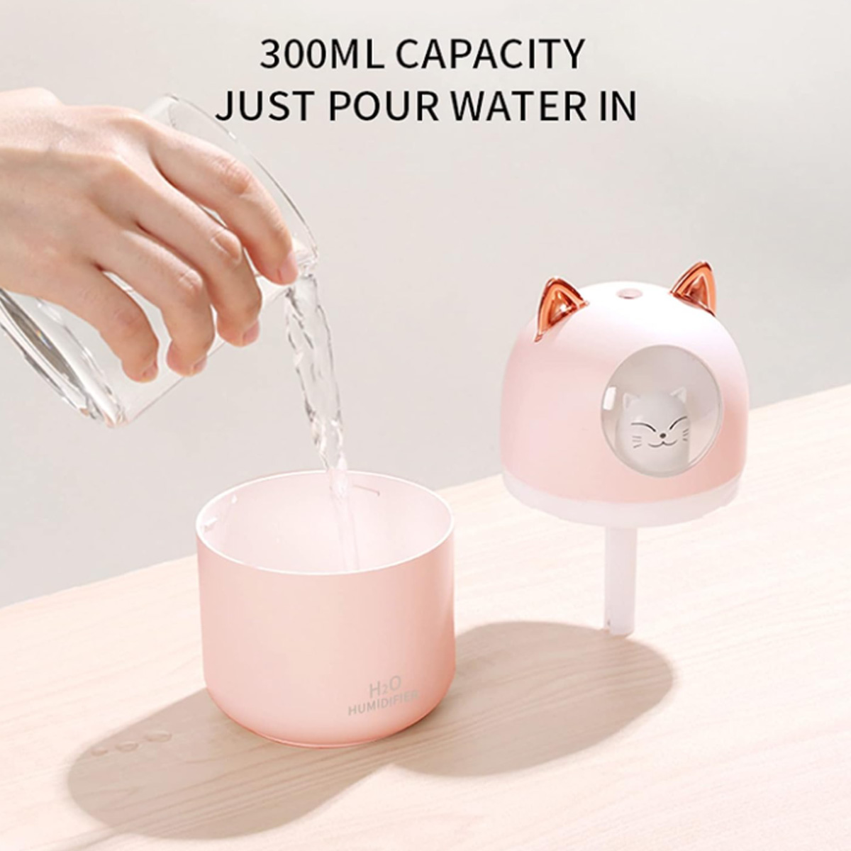 Hand gießt Wasser in einen rosa katzenförmigen Luftbefeuchter. Text sagt 300 ml Fassungsvermögen.