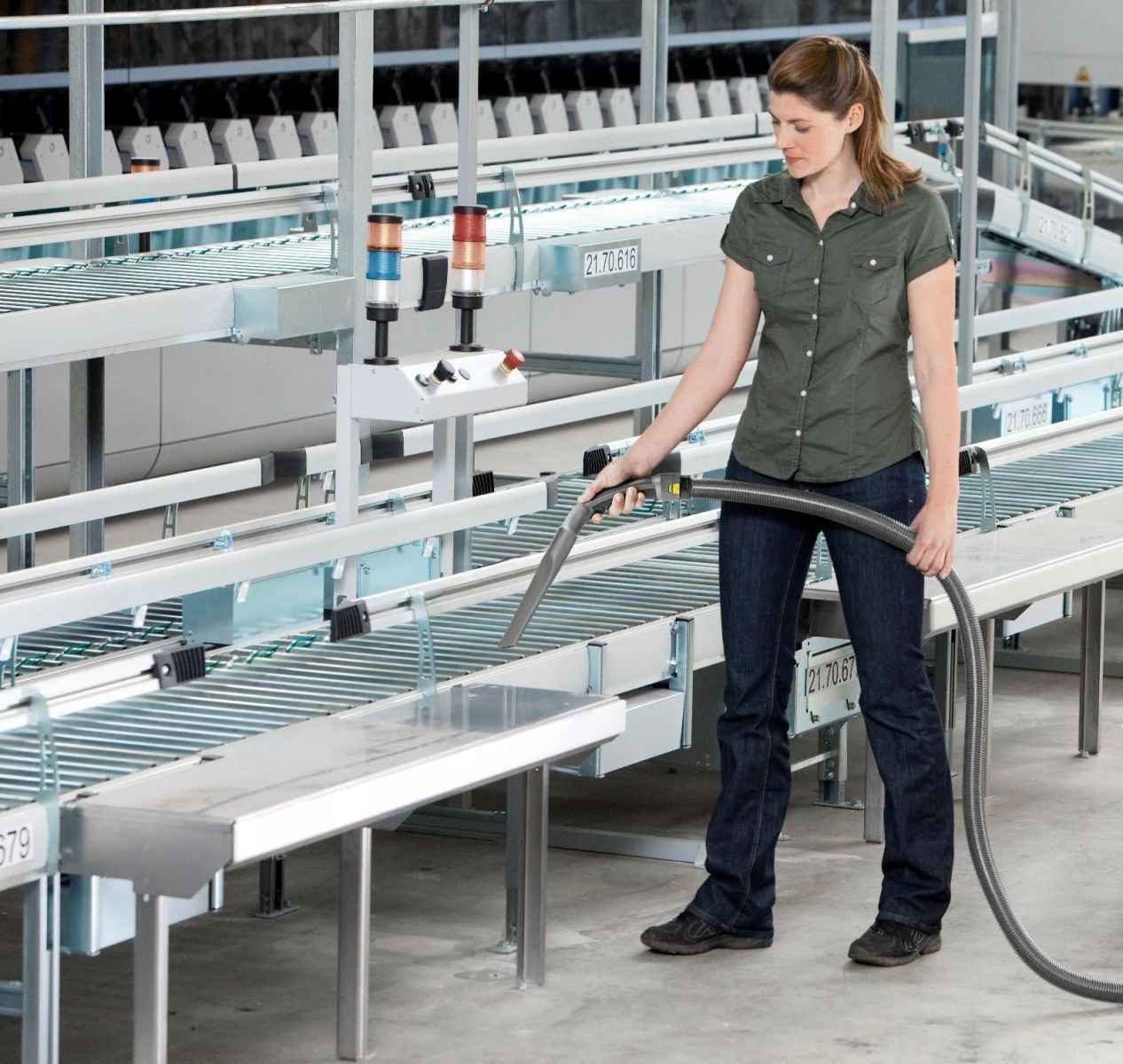 Eine Frau reinigt mit einem Staubsauger ein Förderbandsystem. Industrielle Umgebung mit Metallstrukturen.