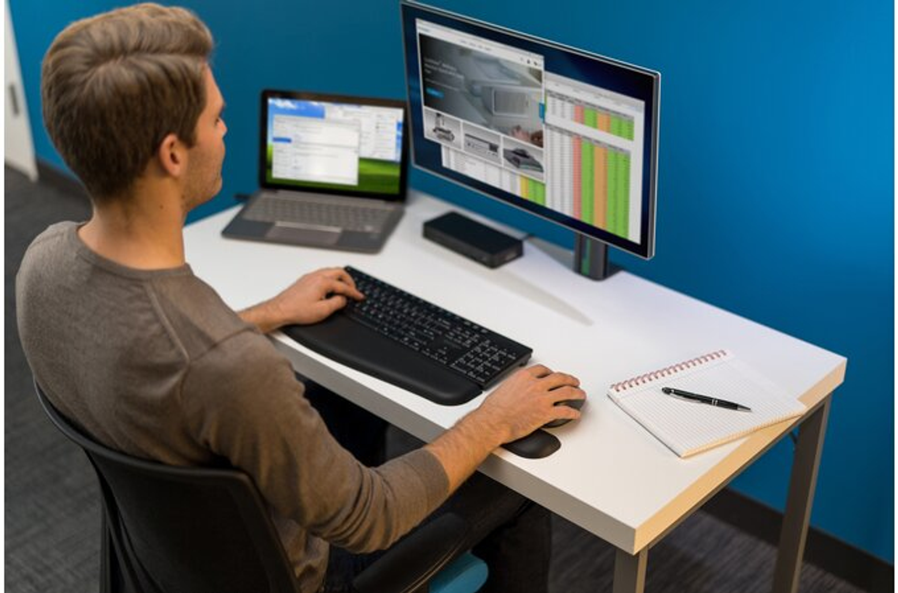 Una persona en un escritorio trabaja con una laptop, monitor, teclado y ratón. Fondo azul.