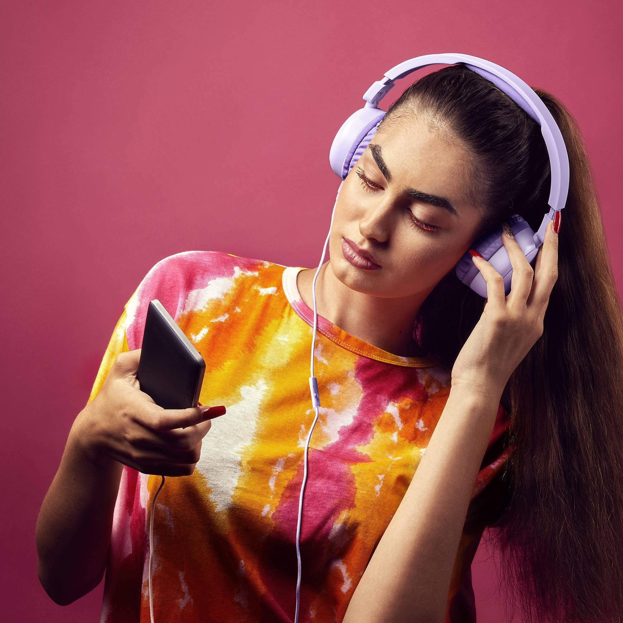 Kobieta z długimi włosami w liliowych słuchawkach, trzymająca telefon i koszulę w stylu tie-dye.