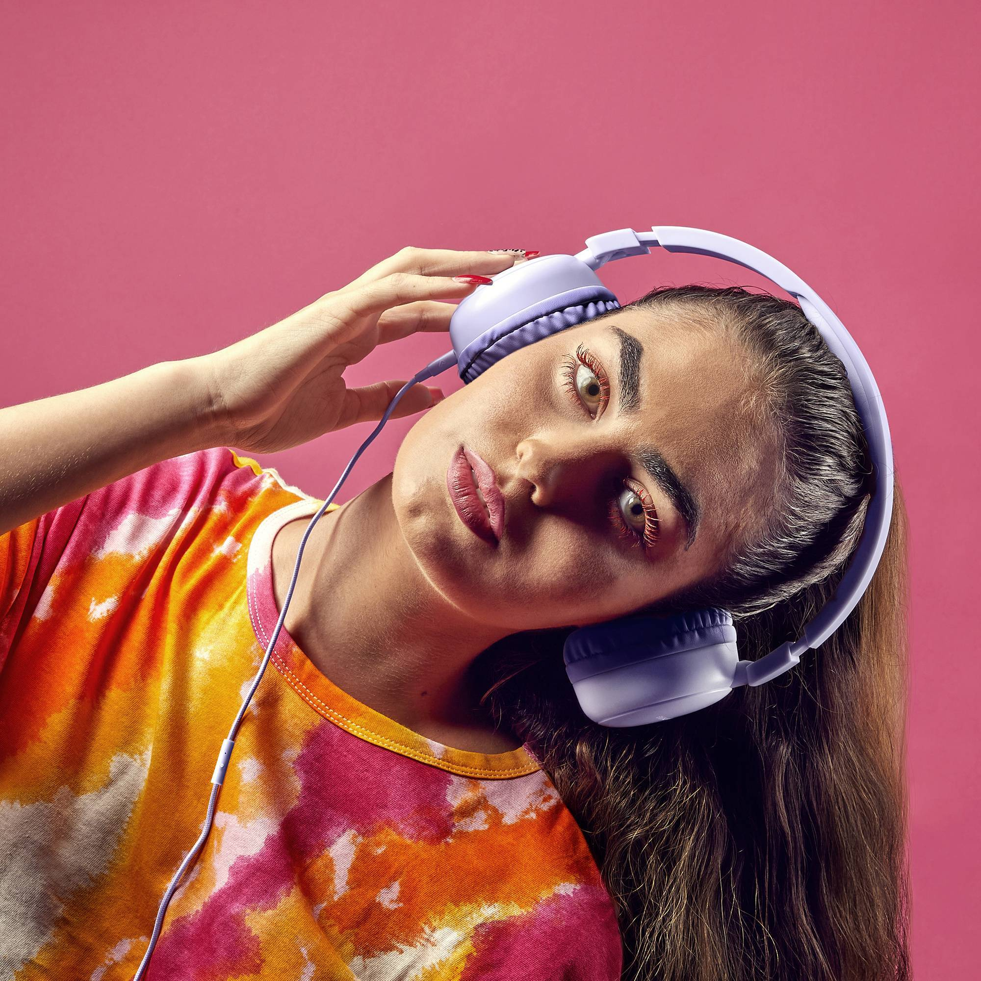 Kobieta w fioletowych słuchawkach. Różowe tło. Patrzy w kamerę.