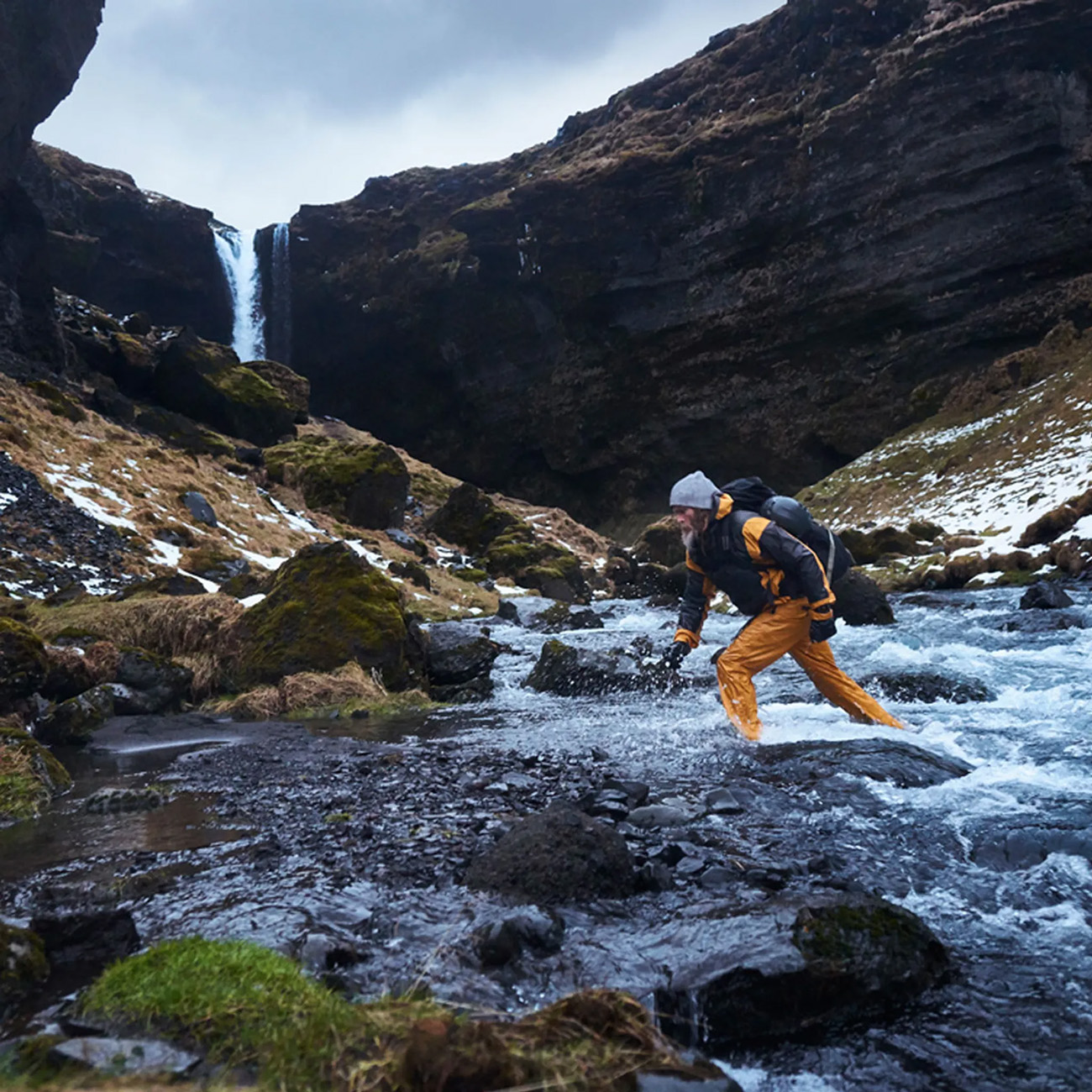 Ein Mann watet in Island durch einen Fluss, in der Nähe eines Wasserfalls.