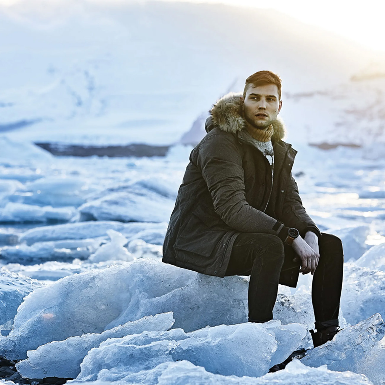 Mann mit Wintermantel mit Pelzkragen, sitzt auf Eis. Hintergrund: schneebedeckter Berg.