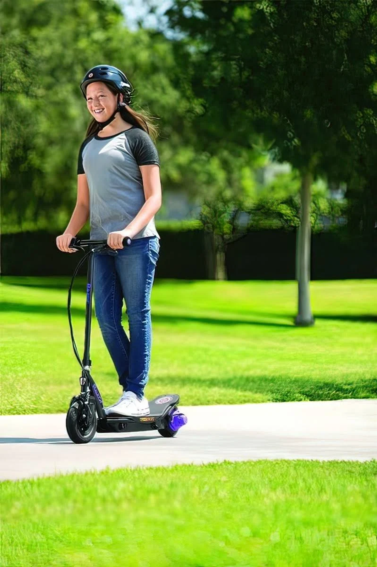 Una giovane donna guida uno scooter elettrico Razor sul marciapiede. Indossa un casco e jeans. Erba verde e alberi sullo sfondo.