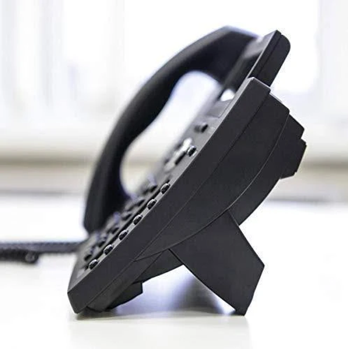 Telefono da scrivania nero su superficie bianca, vista angolata. Pulsanti e cornetta visibili.