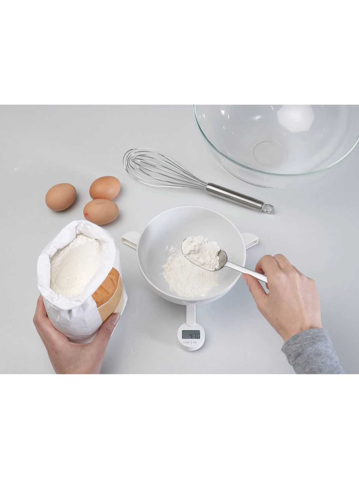 Scena di cucina: farina in una ciotola su bilancia, uova, frusta, sacco farina.