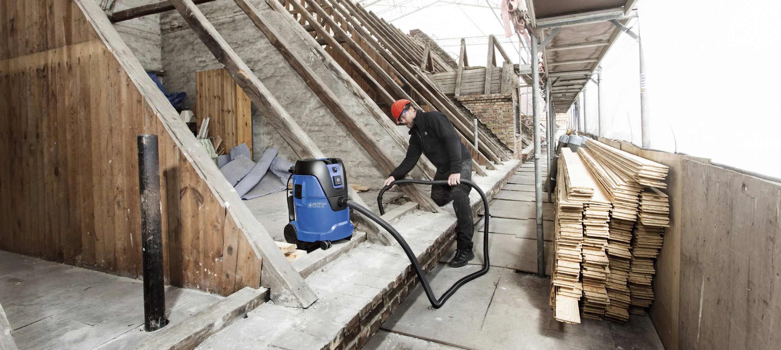 Mann in einer Werkstatt, der einen blauen Staubsauger benutzt, Holzkonstruktion im Hintergrund.