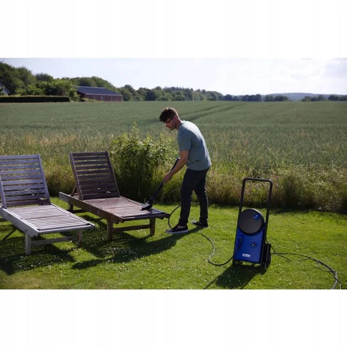 Mężczyzna myje krzesło z drewna myjką ciśnieniową obok drugiego krzesła na polu.