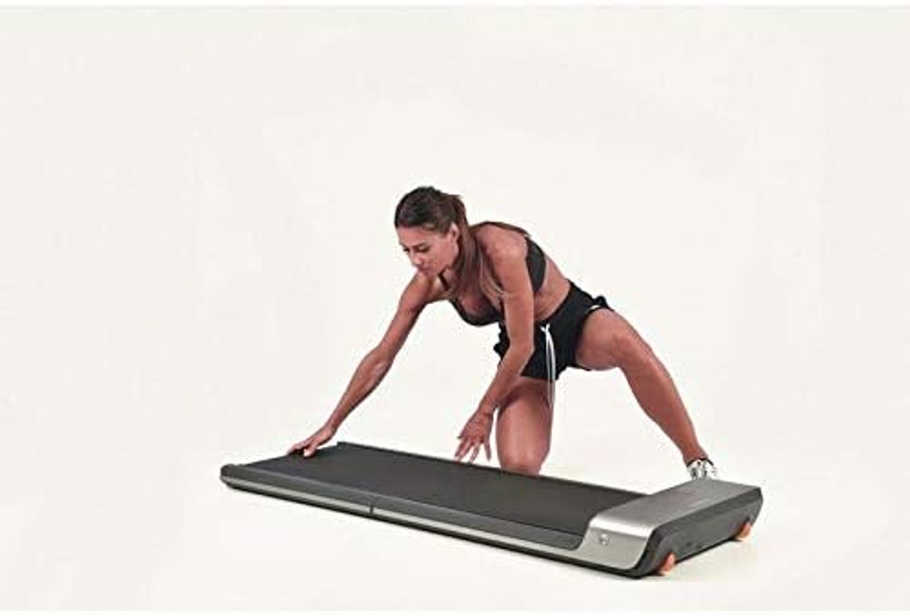 A woman kneels and touches a small treadmill. The treadmill is silver and black, on a white background.