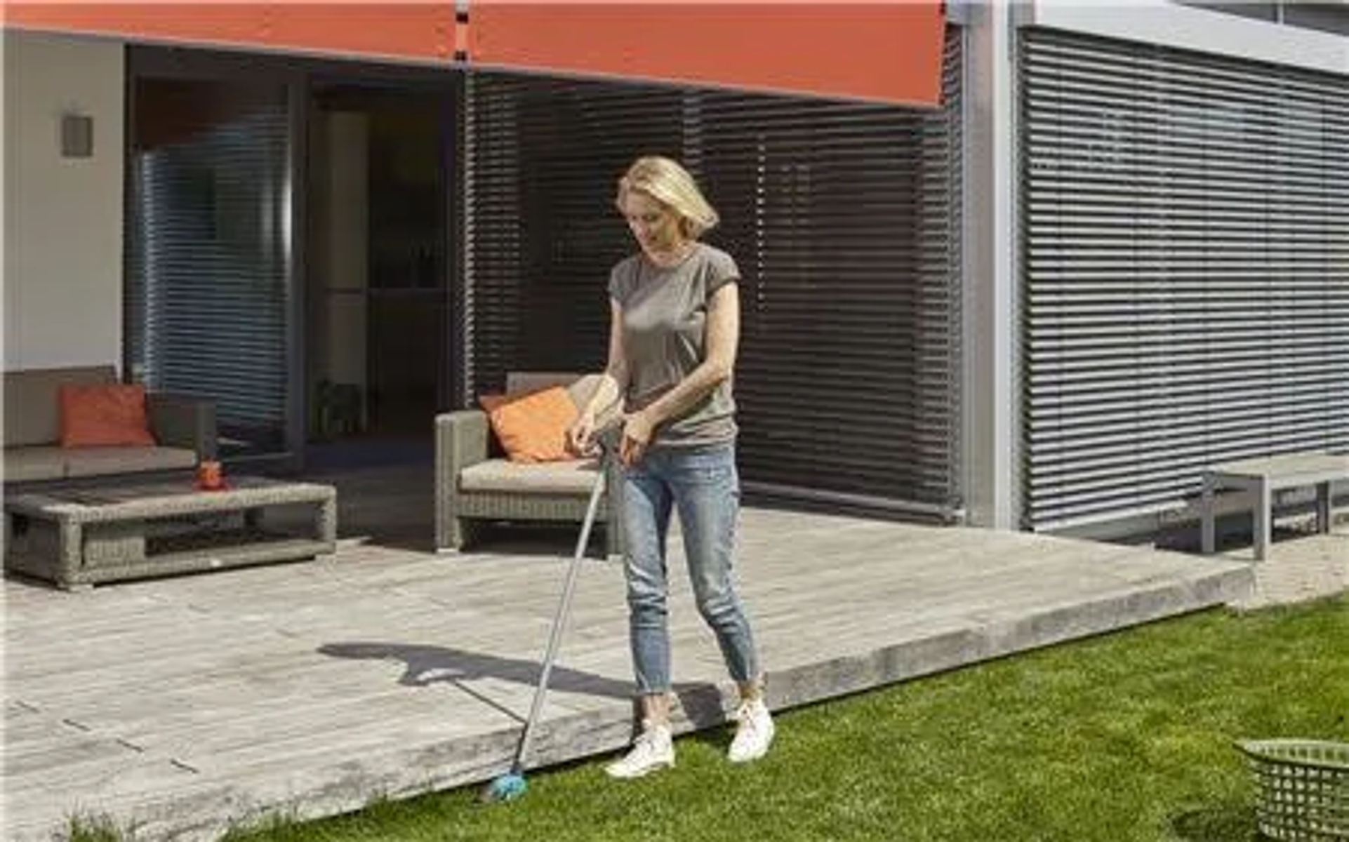 Frau fegt Holzterrasse mit Besen, Gartenmöbel im Hintergrund.