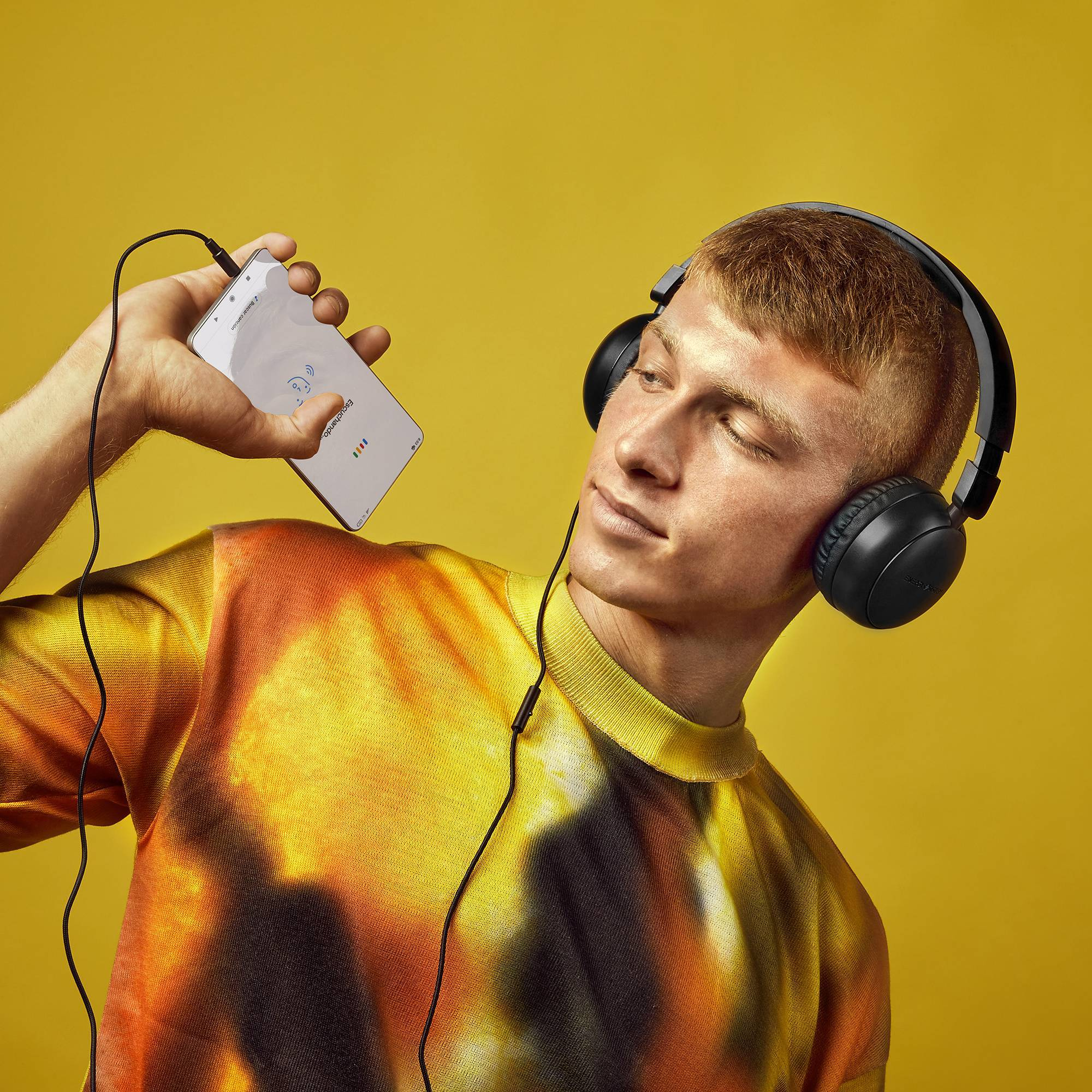 Un uomo con le cuffie tiene in mano un telefono che mostra un segnale wifi. Sfondo giallo.